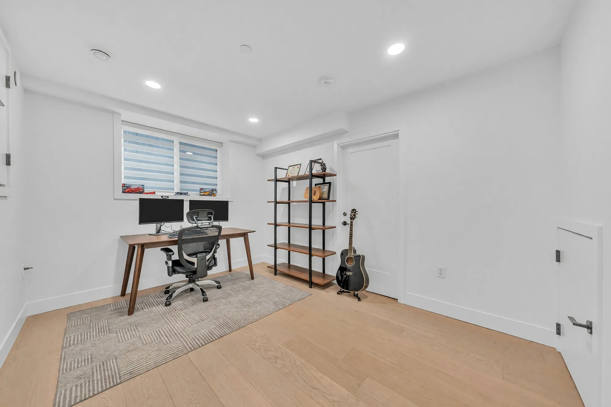 The den: a desk and an office chair, open shelving, a rug, a window with a blind