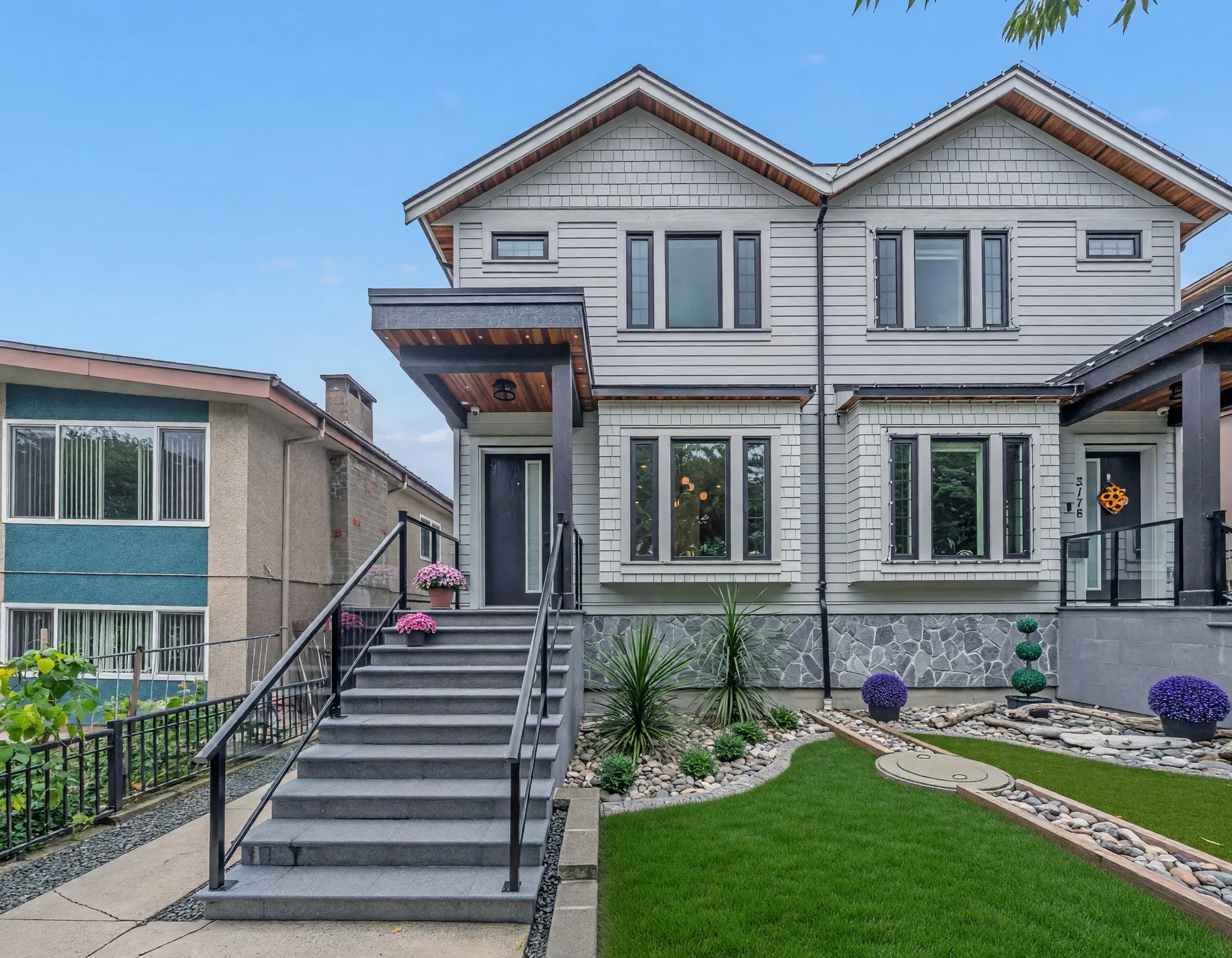 The front of the duplex in daylight: two storeys of pale grey shingle and siding over a stone base, two sets of steps with black railings, lawn either side of a paved strip