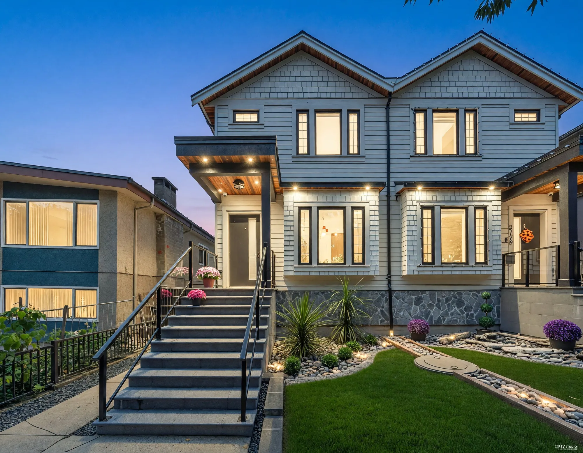3180 Graveley Street from the street at dusk: the left-hand half of a two-storey duplex in pale shingle over stone, the windows and the gate lanterns lit