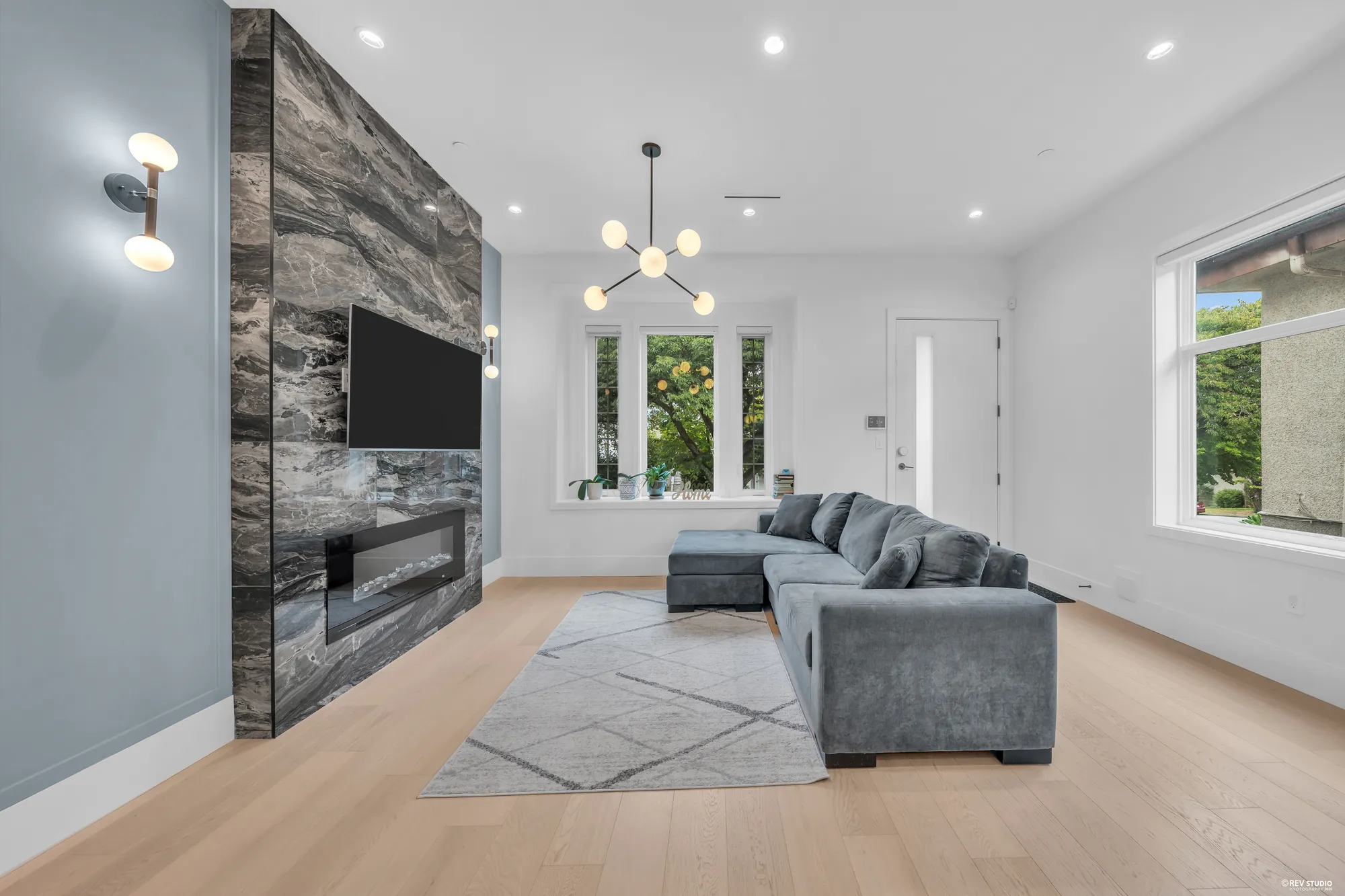 The living room: a grey sectional facing a fireplace set in a dark marbled stone wall with a television above it, the front door and the front windows beyond