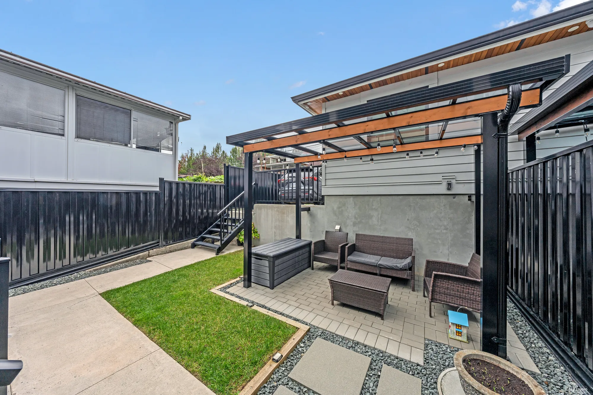The patio at ground level: a pergola over an outdoor sofa set, a strip of lawn beside the paving, black fencing