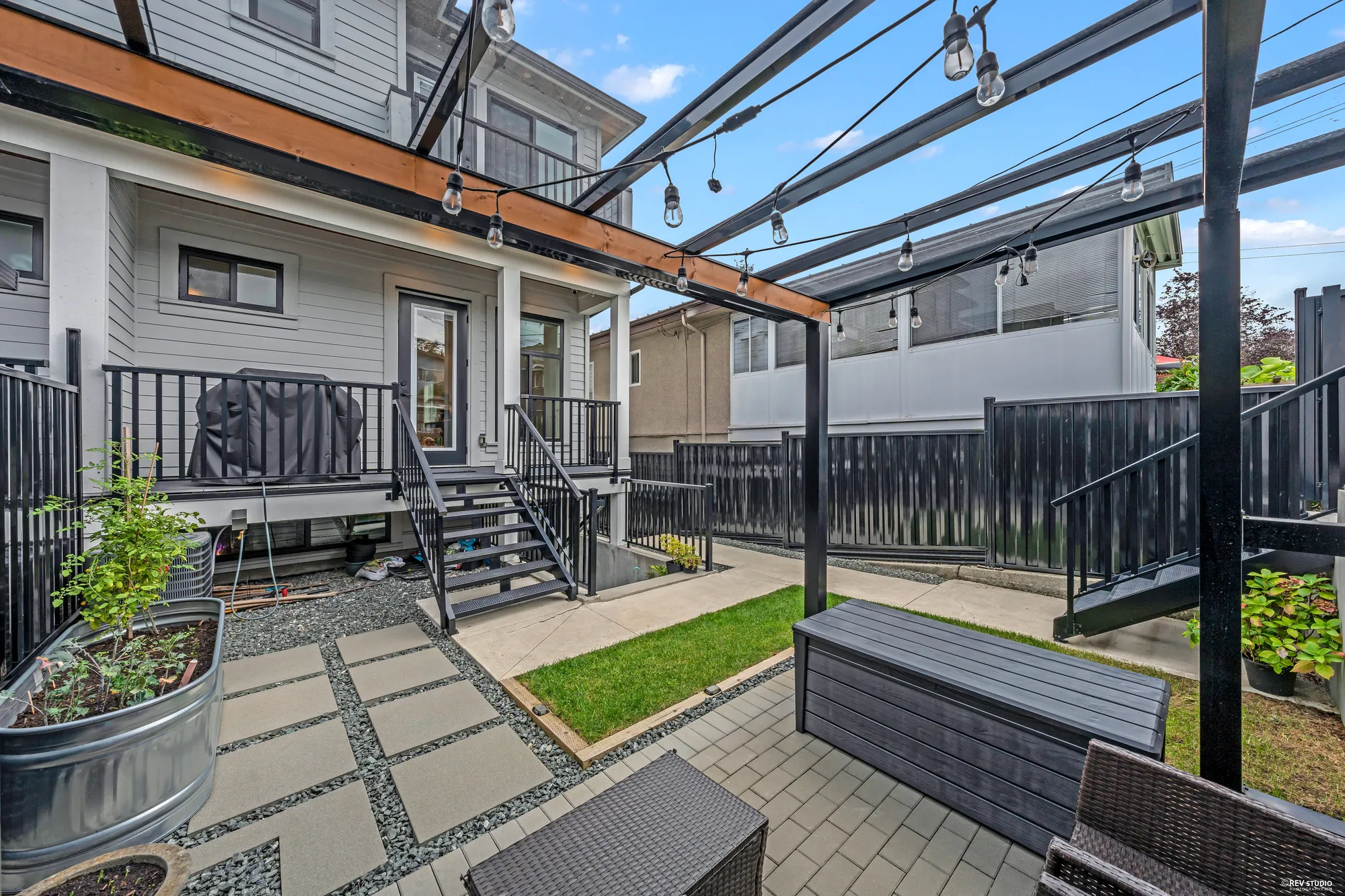 The patio from beside the house, the stairs from the deck coming down to it, string lights overhead