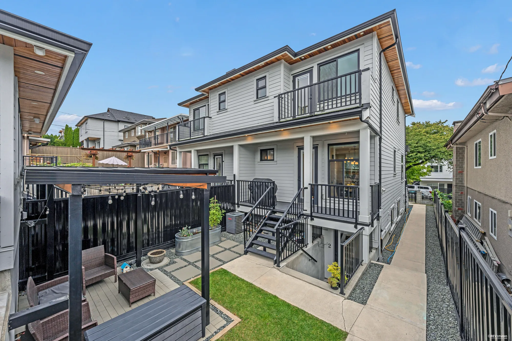 The back of the building from the garage: two storeys of siding with a balcony above a deck, a pergola with string lights over the patio, a strip of lawn and black railings