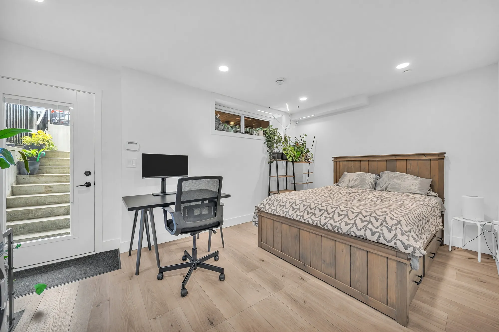 The same room from the kitchen end: a bed under a small high window, a desk, and the door to the side patio with steps rising outside it