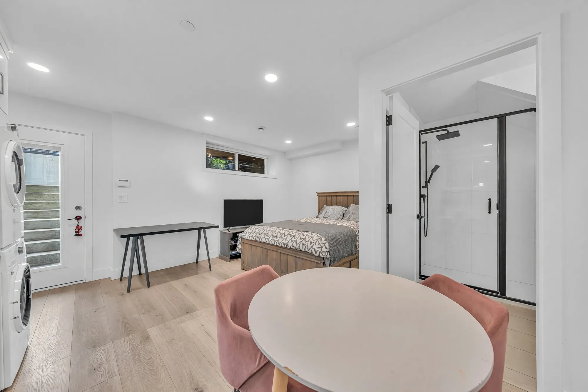 A round table and two pink chairs, the door to the bathroom and its glass shower on the right, the outside door at the far end