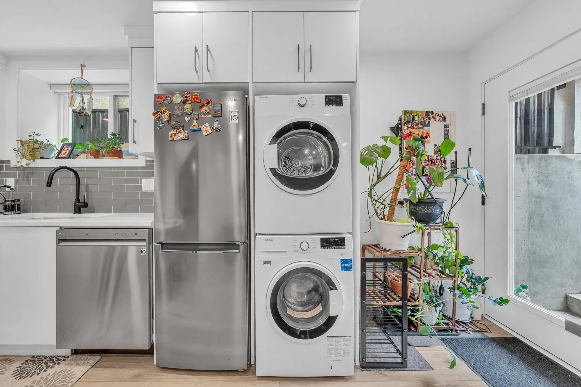 The lower kitchen: white cabinets, a stainless fridge and dishwasher, a stacked washer and dryer, plants on a stand by the door